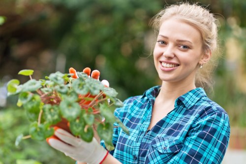 A young woman with blonde hair tied back, wearing a blue and green plaid shirt and white gardening gloves, is standing outdoors in a garden. She is holding a terracotta pot containing a small, lush green plant with rounded leaves and reddish stems. The background features blurred trees and shrubs, indicating a well-maintained outdoor space with natural light and mild weather conditions, suitable for gardening activities. The scene reflects a typical residential garden environment in Newham, London, with neatly arranged plant beds and organic soil visible at the base of the plants, aligning with professional gardening and landscaping services offered by Landscapers Newham.