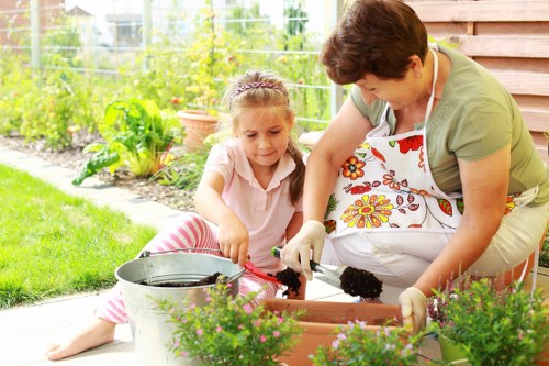 A garden scene in a residential outdoor space shows a woman and a young girl working together on a gardening project. The woman, wearing a white apron with a colorful floral pattern, is kneeling on the paving and handling a small gardening tool or plant, while the girl, dressed in a light pink top and pink-striped leggings, sits nearby on the patio. In front of them, there are flower pots filled with blooming plants, and a metal watering can is positioned on the paving. The garden backdrop features a lush, green lawn, a variety of leafy shrubs, and flower beds with vibrant vegetation, all suggesting a well-maintained, landscaped yard typical of a suburb in Newham. The weather appears bright and clear, with natural sunlight illuminating the scene, highlighting the greenery and the natural tones of the plants and garden surfaces. This setup exemplifies professional gardening and planting services, similarly offered by landscapers in Newham or surrounding areas, aiming to create inviting and flourishing outdoor spaces.