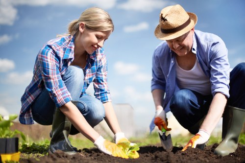In a bright outdoor garden setting, two individuals are engaged in planting activities on a freshly tilled garden bed with rich, dark soil. The younger woman, dressed in a plaid shirt and jeans, is carefully placing a seedling or small plant into the ground, while the older woman, wearing a hat, light jacket, and gloves, is using a trowel to assist with the planting process. The background reveals a clear, blue sky with some clouds, and surrounding elements include low-growing green plants and the edge of a lawn area. The scene captures a moment of outdoor gardening, emphasizing soil preparation and planting, typical of garden care and maintenance services offered by local landscapers such as Landscapers Newham. The natural light highlights the vibrant green of the plants and the textures of the soil, creating a realistic and inviting image that underscores the importance of garden cultivation and outdoor landscape improvement around Newham.