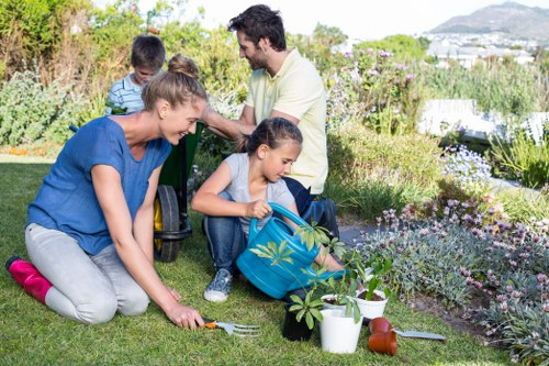 A family of four engaging in gardening activities in a well-maintained backyard garden. The scene shows two children, a girl and a boy, planting or tending to potted plants placed on the grass alongside gardening tools such as a small shovel and a watering can. An adult woman is kneeling nearby, assisting or observing the children, wearing a blue top and grey trousers, while an adult man stands slightly behind, supervising and smiling at the activity. The garden features a lush, green lawn with a neat border of flowering plants and shrubs, bordered by a stone edge. In the background, there are taller bushes and trees, with a view of the sky and distant landscape, indicating an outdoor, suburban environment typical of Newham. The scene is illuminated by natural daylight, suggesting clear weather suitable for outdoor gardening. This image exemplifies family outdoor activities and garden maintenance services that a professional landscaper such as Landscapers Newham might support, including planting, lawn care, and general garden upkeep in residential outdoor spaces near the postcode area of E13.