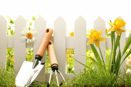 A close-up of a neatly maintained garden border in a residential outdoor space, featuring a white picket fence in the background. In the foreground, vibrant yellow daffodils with long green leaves grow alongside patches of lush, dense grass that appears healthy and well-kept. Next to the flower bed, there are two gardening tools—one with a wooden handle and a small trowel, and another with a similar wooden handle, possibly a cultivator—resting on the soil and grass. The garden area shows signs of recent planting or maintenance, with bright natural lighting indicating a clear and sunny day. The overall scene captures a well-organized garden space typical of residential properties in Newham, highlighting elements of lawn care and gardening that Local landscapers like Landscapers Newham may service. This image emphasizes outdoor upkeep on a tidy and inviting garden, incorporating natural tones and textures suited for professional lawn and garden care in the local area.