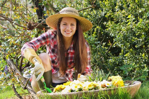 A young woman with long brown hair, wearing a wide-brimmed straw hat, a red and white checkered shirt, beige gardening gloves, and light-colored trousers, is kneeling in a lush garden. She is smiling at the camera while tending to a flower bed filled with blooming yellow and white flowers. Behind her, there are various green shrubs, leafy trees, and a partly shaded outdoor space, indicating a well-maintained backyard or front garden area in Newham. The garden includes a neatly edged grassy lawn, and the flowering bed is bordered by natural soil and mulch. Sunlight filters through the foliage, creating a bright and inviting atmosphere suitable for outdoor gardening activities. The scene reflects active gardening work, aligned with landscaping services provided by companies like Landscapers Newham, with viewable elements emphasizing garden upkeep and outdoor planting in a suburban environment.