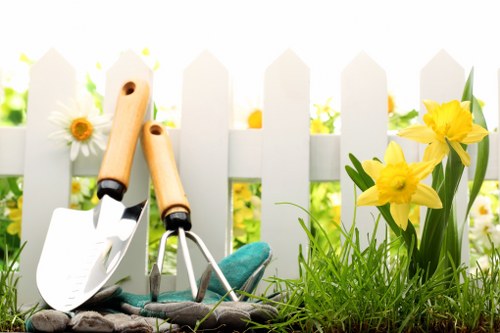 The image depicts a small section of a garden with a white picket fence in the background, situated in Newham. In the foreground, there is a patch of green grass and soil, with a few yellow daffodil flowers in bloom on the right side. Leaning against the fence are garden tools including a trowel and a hand rake, featuring wooden handles and metal heads, positioned on top of a pair of gardening gloves. The scene is illuminated by natural sunlight, suggesting a bright, clear day, and the overall setting presents a well-maintained outdoor space suitable for gardening activities. This setup reflects typical elements of a residential garden in London, with potential for landscaping or lawn care services by companies such as Landscapers Newham, emphasizing the importance of outdoor maintenance and garden enhancement in local communities.
