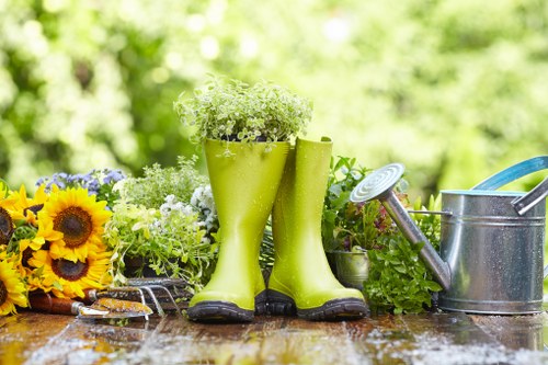 The image displays a garden scene with a wooden outdoor table featuring gardening tools and decorative elements. Prominently, there are a pair of bright green rubber boots filled with white flowering plants, positioned centrally. To the right of the boots, a metal watering can with a long spout is visible, alongside a blue-handled garden tool. On the left side, a bouquet of vibrant yellow sunflowers and various green foliage add colour and texture to the arrangement. The background shows a blurred garden environment with lush green trees and shrubs, illuminated by natural daylight, suggesting a sunny, outdoor setting typical of a garden in Newham, East London. The scene reflects a well-maintained outdoor space suitable for gardening and lawn care services provided by companies like Landscapers Newham, adding natural tones and textures commonly associated with quality outdoor landscaping and garden maintenance.