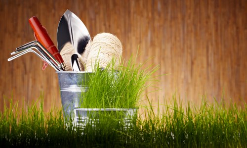 A close-up image of gardening tools and supplies arranged in a small galvanized metal bucket placed on a vibrant, green grass lawn. The tools include a hand trowel with a wooden handle, a three-pronged cultivator, and a small gardening fork, all with metallic heads. Behind the bucket, there is a ball of twine, suggesting general gardening or planting activities. The grass appears healthy and lush, with varying shades of green, indicating a well-maintained outdoor space. The background features a wooden fence in warm, natural tones, creating a cozy garden atmosphere. The overall scene suggests preparation for gardening tasks such as planting, soil tending, or general outdoor maintenance, fitting for a landscaping or gardening service context in Newham or nearby areas.