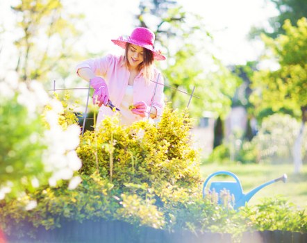 A woman in a pink sunhat and gardening gloves is tending to a vibrant flower bed in a well-maintained garden, with lush green trees and a clear sky in the background. The flower bed features a variety of yellow and white flowering plants, bordered by a low edge and situated on a neatly kept grassy lawn. A blue watering can is visible in the foreground, indicating ongoing garden care. The garden area includes paved pathways and wooden decking, creating an organized outdoor space suitable for gardening activities. The scene is illuminated by natural daylight, suggesting a bright, sunny day. This image exemplifies typical landscaping and gardening features found in residential gardens in Newham, aligning with professional gardening and lawn care services offered by landscapers such as Landscapers Newham. The overall environment is tidy, lush, and inviting, emphasizing the importance of outdoor maintenance and plant care. Natural tones of green, yellow, and pink dominate the scene, providing a vibrant yet harmonious outdoor atmosphere.