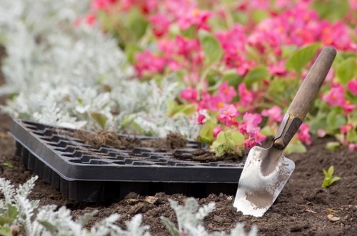 A close-up view of a garden bed in a landscaped outdoor space within Newham, showing freshly dug soil with a small hand trowel with a wooden handle partially inserted into the earth. Behind the soil, there are vibrant pink and white flowering plants, along with green foliage, creating a colourful and well-maintained garden scene. An empty black seed tray with gently overflowing soil rests on the ground, indicating recent planting or soil preparation activities. The environment appears to be part of a carefully landscaped backyard or front garden area, with natural light suggesting a clear day, suitable for gardening and lawn care services offered by Landscapers Newham.