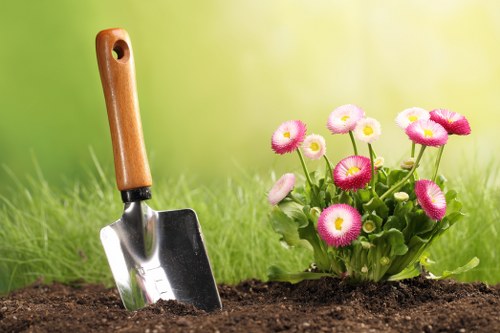 A garden scene featuring a close-up view of freshly turned dark soil with a small gardening trowel inserted upright into the earth, its wooden handle partially visible. To the right, there is a vibrant cluster of pink and white primrose flowers with green leaves, positioned on a neatly maintained lawn area. The background shows a softly blurred green hedge or grass, indicating an outdoor garden space in Newham, with natural daylight illuminating the scene. The overall setting demonstrates typical gardening activity suitable for landscaping or lawn care services, with attention to plant beds and outdoor environment maintenance.