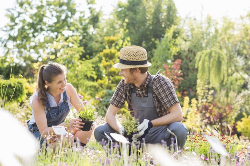 A gardener and a woman are working together in a well-maintained outdoor garden, surrounded by lush green trees and vibrant flowering plants. The gardener, wearing a straw hat and gardening gloves, is tending to a small potted plant while sitting on the grass. The woman, with long dark hair tied back, is smiling and holding a similar potted plant, appearing engaged in the gardening activity. The garden features a mixture of flower beds with blooming flowers in various colours, neatly edged borders, a lush green lawn, and some shrubs in the background. The scene is illuminated by natural daylight, suggesting fair weather with soft sunlight filtering through the trees, creating a serene and inviting outdoor atmosphere consistent with professional gardening and landscaping efforts in the Newham area.