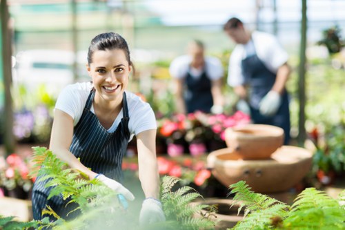 A young woman wearing a striped apron and gardening gloves is smiling at the camera while tending to lush green ferns in a bright, well-lit greenhouse or garden centre environment. Behind her, there are blurred figures of another person and various potted plants, including flowering plants with pink blooms, indicating a horticultural setting. The scene features a mixture of vibrant green foliage, some textured soil beds, and decorative stone water features in the background, suggesting a focus on plant nurturing and garden landscaping. The natural daylight streaming through the transparent roof emphasizes the clarity and health of the plants, and the overall environment appears tidy and professionally maintained, aligned with gardening and landscaping services that create attractive outdoor spaces in Newham or nearby areas. This detailed scene highlights aspects of plant care, outdoor garden maintenance, and landscape design that a company like Landscapers Newham might support through their gardening services.